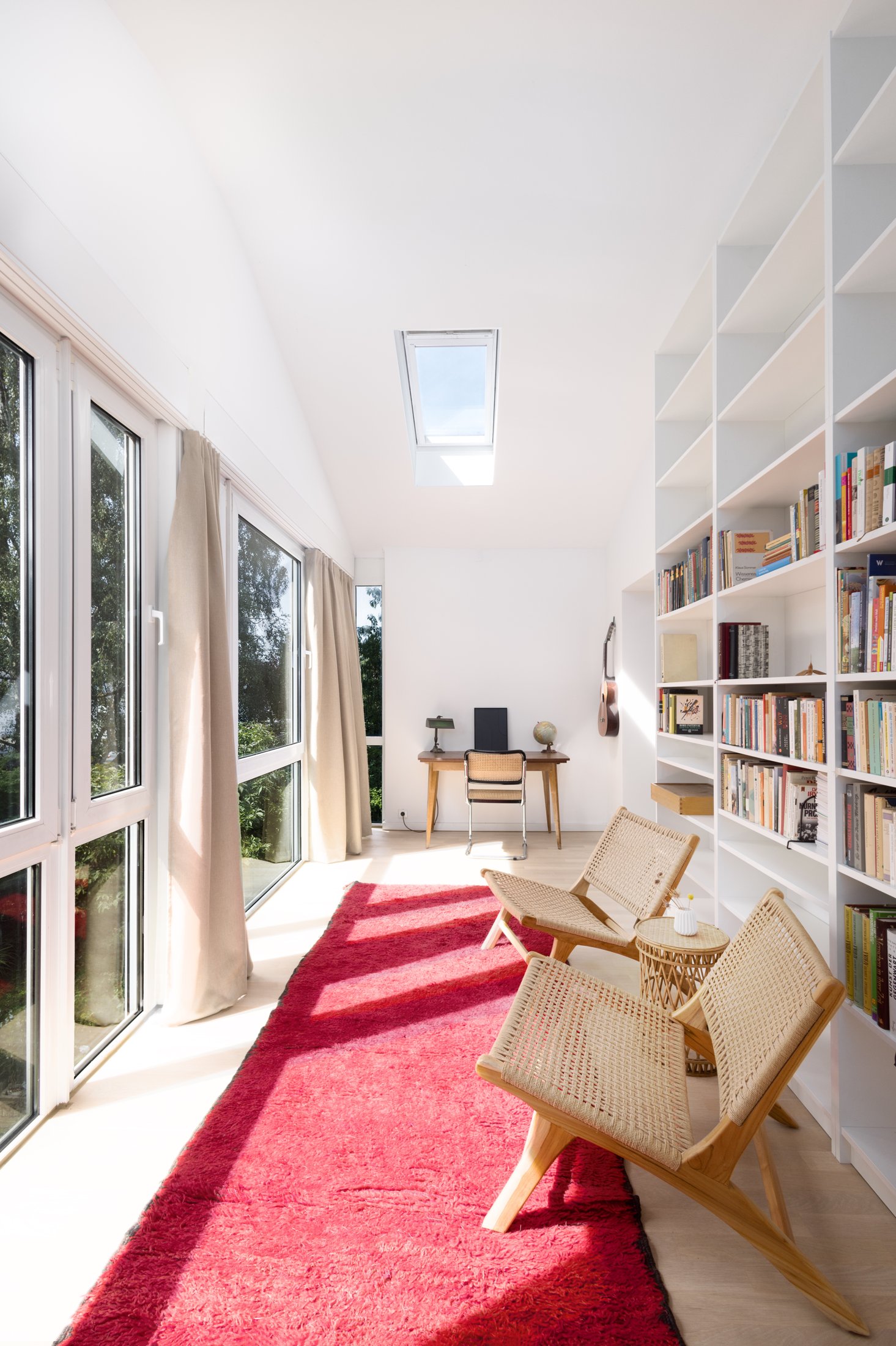 Projekt Taubenweg: Schmaler Raum mit Schreibtisch, Flechtstühlen, rotem Teppich und Bücherregal