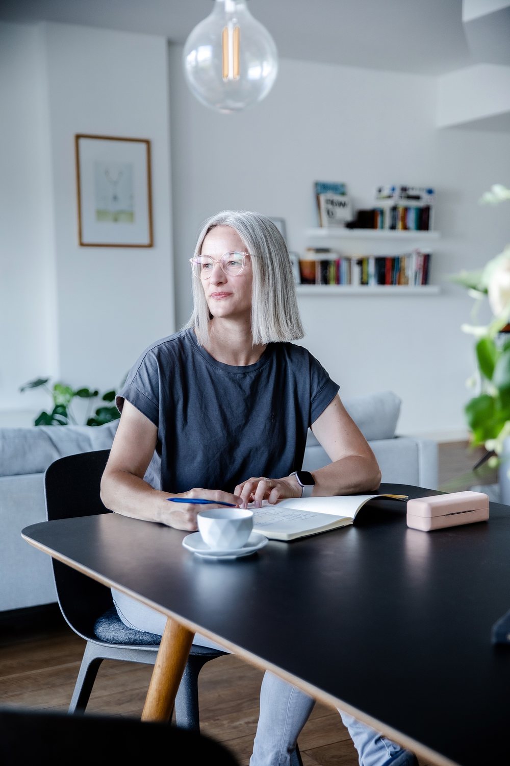 Businessporträt: Frau mit Brille am Tisch mit Notizbuch und Kaffeetasse, Blick aus dem Fenster