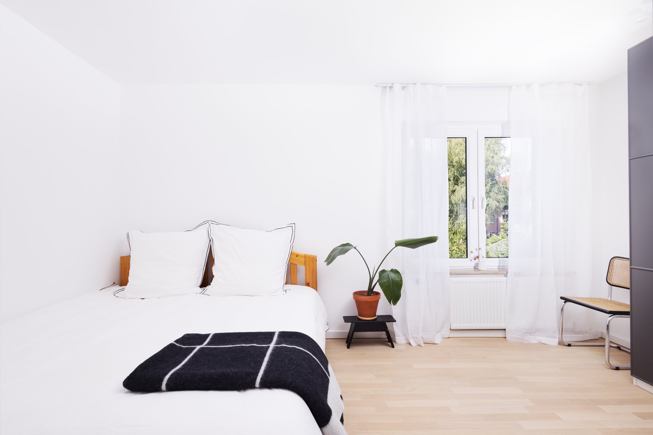 Projekt Taubenweg: Helles Schlafzimmer mit weißer Bettwäsche, schwarzer Decke und Zimmerpflanze