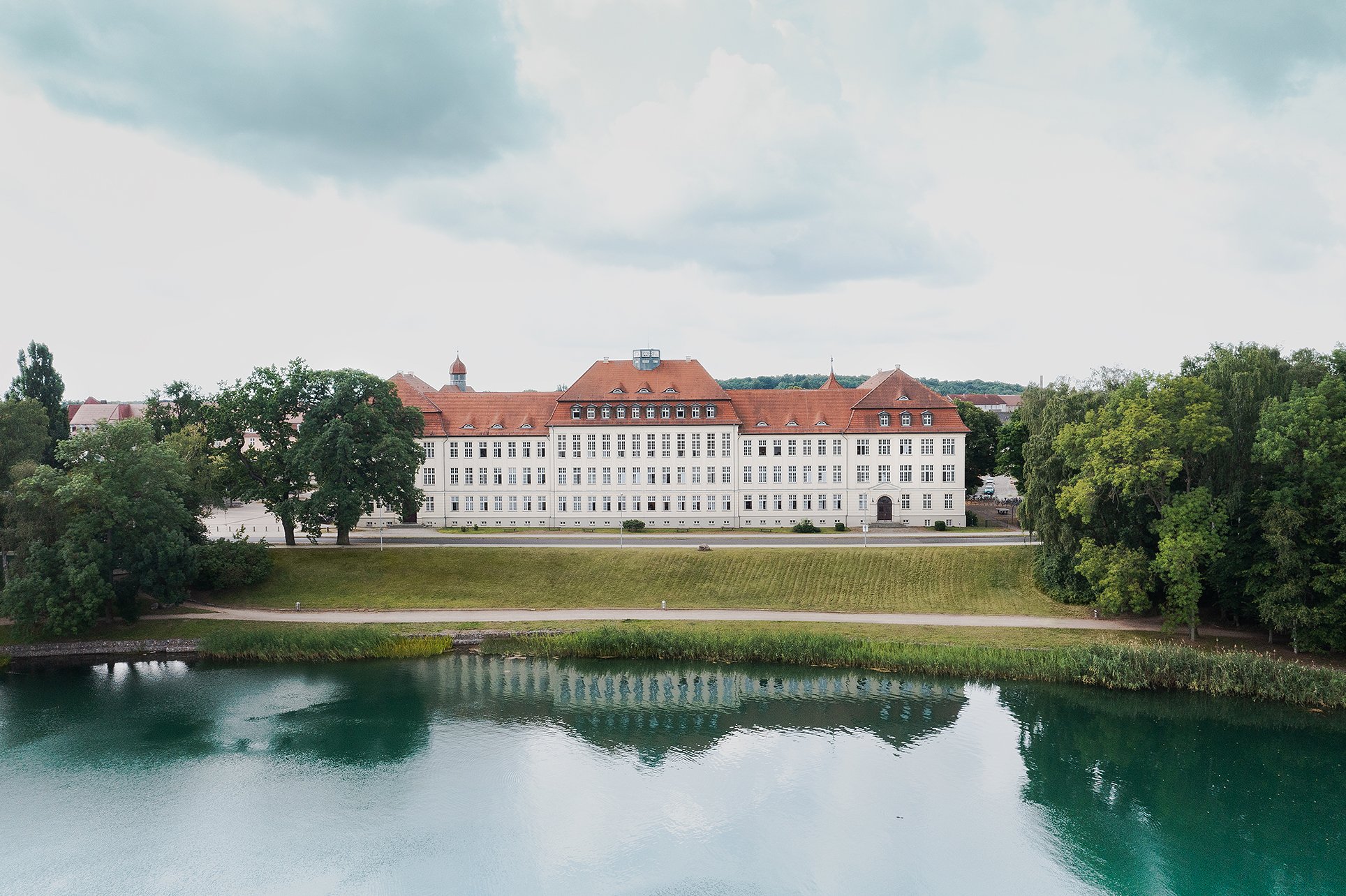 Drohnenaufnahme: historisches Schulgebäude mit rotem Ziegeldach hinter einer Wiese, im Vordergrund ein See, Projekt DENA