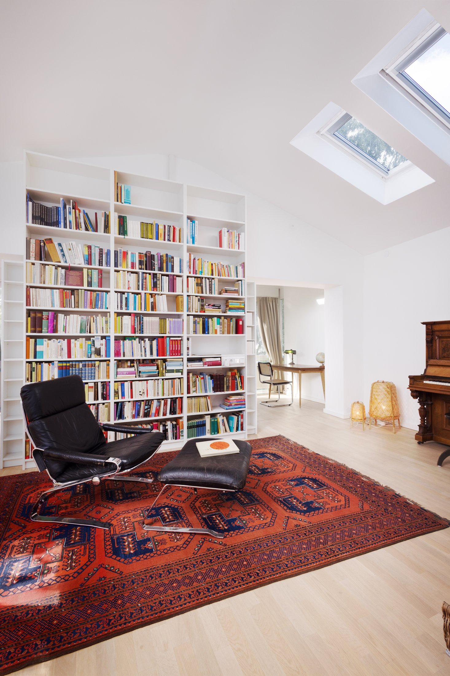Projekt Taubenweg: Bibliothek mit hohem Bücherregal, Ledersessel und rotem Teppich unter Dachfenstern